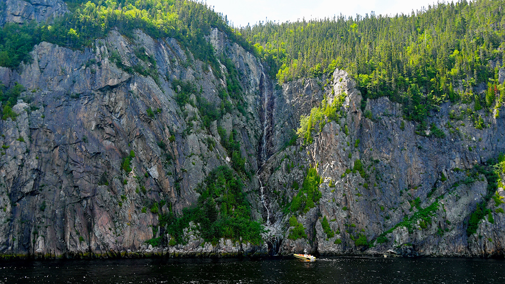 A narrow waterfall flows down a rocky cliff covered in dense vegetation. At the base of the cliff, a small yellow boat floats on dark water, highlighting the grandeur of the rocky wall and the tranquility of the surrounding nature.