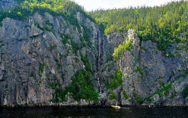 A narrow waterfall flows down a rocky cliff covered in dense vegetation. At the base of the cliff, a small yellow boat floats on dark water, highlighting the grandeur of the rocky wall and the tranquility of the surrounding nature.