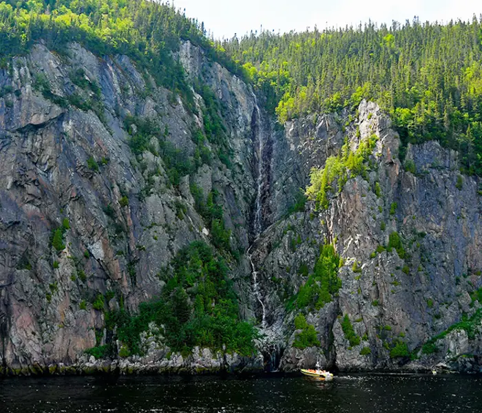 A narrow waterfall flows down a rocky cliff covered in dense vegetation. At the base of the cliff, a small yellow boat floats on dark water, highlighting the grandeur of the rocky wall and the tranquility of the surrounding nature.