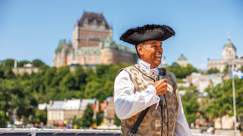 Un joyeux guide touristique en costume historique parle dans un microphone avec le Château Frontenac en arrière plan par une journée ensoleillée.