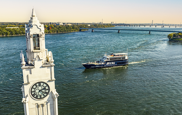Le navire AML Cavalier Maxim naviguant sur le fleuve Saint-Laurent avec au premier plan la tour de l'horloge de Montréal et en arrière plan l'Ile Sainte Hélène et le Pont Samuel de Champlain avec une lumière dorée.