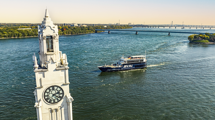 The AML Cavalier Maxim ship sailing on the Saint Lawrence River with the Montreal Clock Tower in the foreground and the Saint Helen's Island and the Samuel de Champlain Bridge in the background with a golden light.