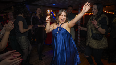 A woman in a blue dress joyfully holding a sparkler in the air, celebrating Christmas surrounded by friends.