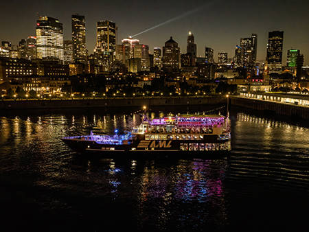 An illuminated "AML" cruise ship sails on a river at night, with a stunning view of skyscrapers and city lights in the background.