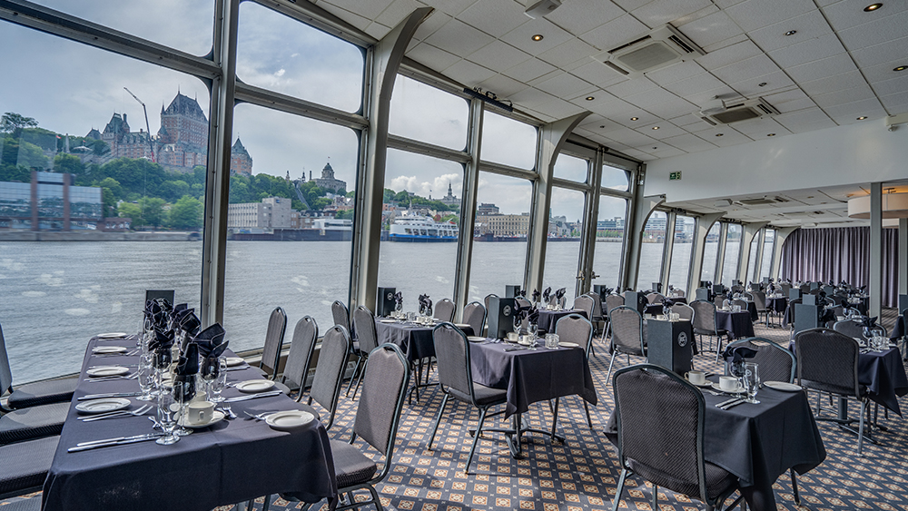 Salle intérieure avec des tables dressées devant une fenêtre donnant vue sur le fleuve Saint-Laurent, le Château Frontenac et le cap Diamant à Québec en arrière plan 