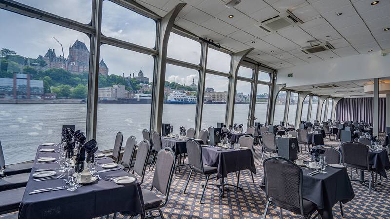 Indoor room with set tables in front of a window overlooking the Saint Lawrence River, the Château Frontenac, and Cap Diamant in Quebec City in the background