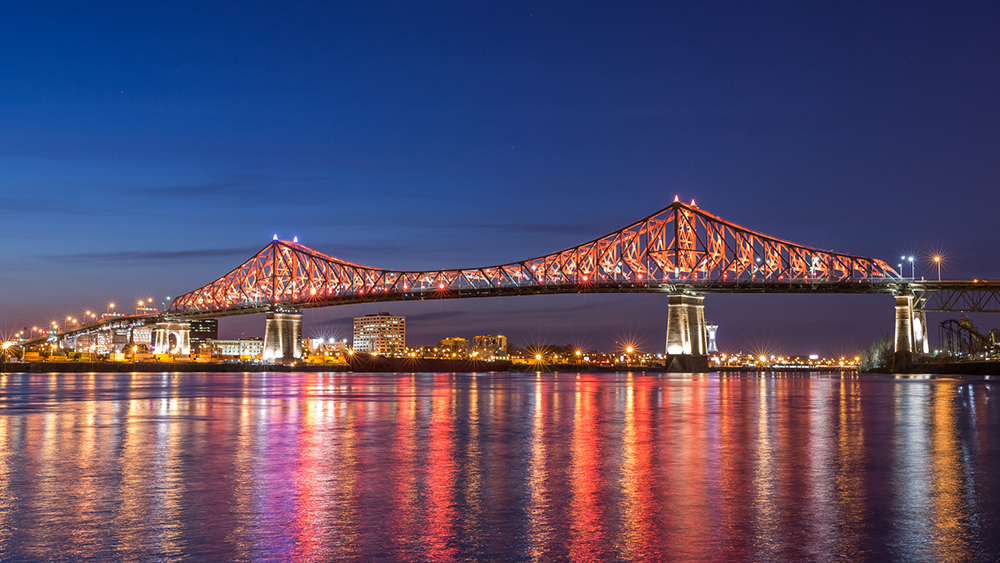 The Jacques-Cartier Bridge lit up in red and white, reflecting on a calm river at night, with city lights and a deep blue sky in the background.