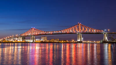 The Jacques-Cartier Bridge lit up in red and white, reflecting on a calm river at night, with city lights and a deep blue sky in the background.