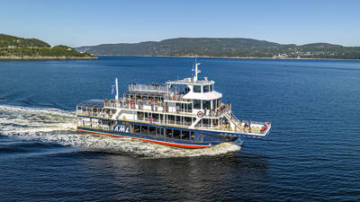 A tour boat cruises on calm water, with passengers on board enjoying the view. The Canadian flag waves at the back of the boat, and green hills and shores are visible on the horizon under a clear sky.