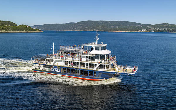 A tour boat cruises on calm water, with passengers on board enjoying the view. The Canadian flag waves at the back of the boat, and green hills and shores are visible on the horizon under a clear sky.