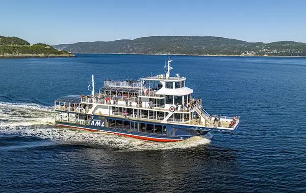 A tour boat cruises on calm water, with passengers on board enjoying the view. The Canadian flag waves at the back of the boat, and green hills and shores are visible on the horizon under a clear sky.