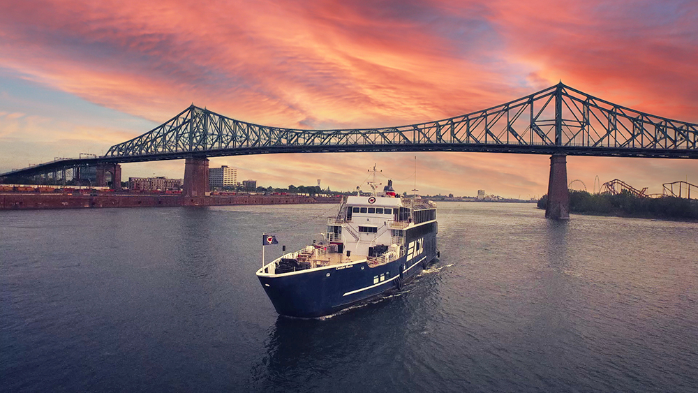 A blue cruise ship labeled "AML" sails on a river at sunset, with the Jacques-Cartier Bridge in the background under a stunning sky with pink and orange hues.