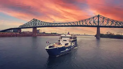 A blue cruise ship labeled "AML" sails on a river at sunset, with the Jacques-Cartier Bridge in the background under a stunning sky with pink and orange hues.