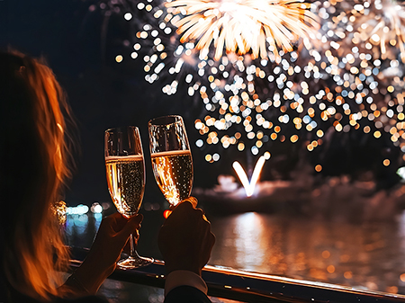Deux personnes trinquent avec des coupes de champagne sur un balcon, admirant un feu d&#x27;artifice brillant illuminant le ciel nocturne et se refl&#xE9;tant sur l&#x27;eau.