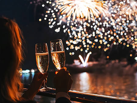 Two people clink champagne glasses on a balcony, enjoying a dazzling fireworks display lighting up the night sky and reflecting on the water.