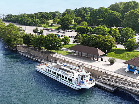 The ship "Tandem" is docked along a quay surrounded by greenery. Trees, a pedestrian pathway, buildings with sloped roofs, and a nearby parking lot are visible. The water is calm, and the scene takes place on a sunny day.