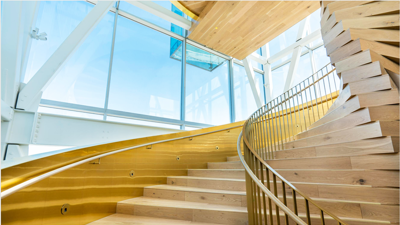 A modern wooden staircase with a gentle curve ascends alongside glass panels, illuminated by natural light in the contemporary architectural setting of the Montreal Port Tower.