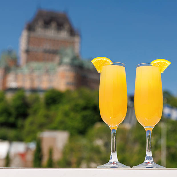 Deux cocktails mimosas, garnis de tranches d'orange sur le bord du verre, sont placés au premier plan. En arrière-plan flou, on distingue un bâtiment historique orné, avec un toit vert et un ciel bleu au-dessus.