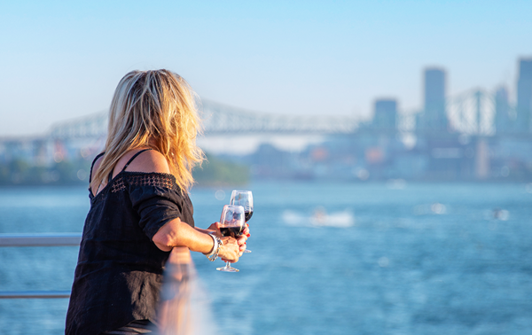 A woman holding a glass of red wine on one of the terraces of the AML Cavalier Maxim ship sailing on the St. Lawrence River, with a view of the Montreal skyline in the background.