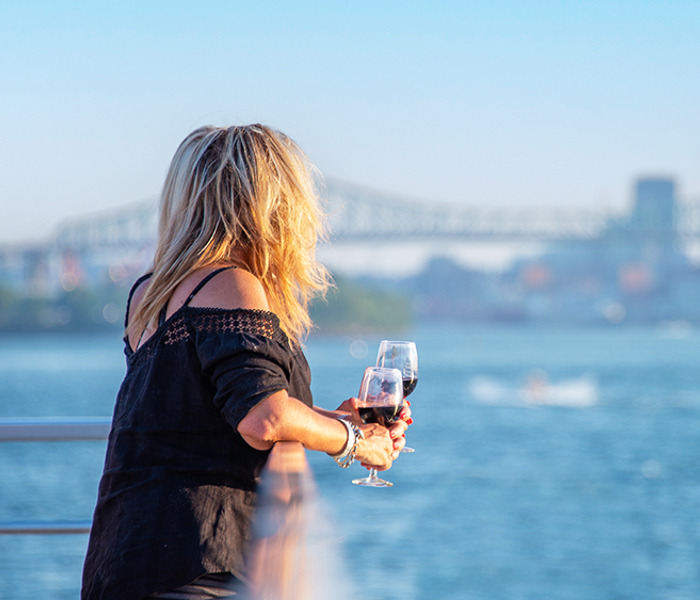 Une femme tient un verre de vin rouge sur l'une des terrasses du navire AML Cavalier Maxim naviguant sur le fleuve Saint-Laurent avec une vue sur la skyline de Montréal en arrière plan. 