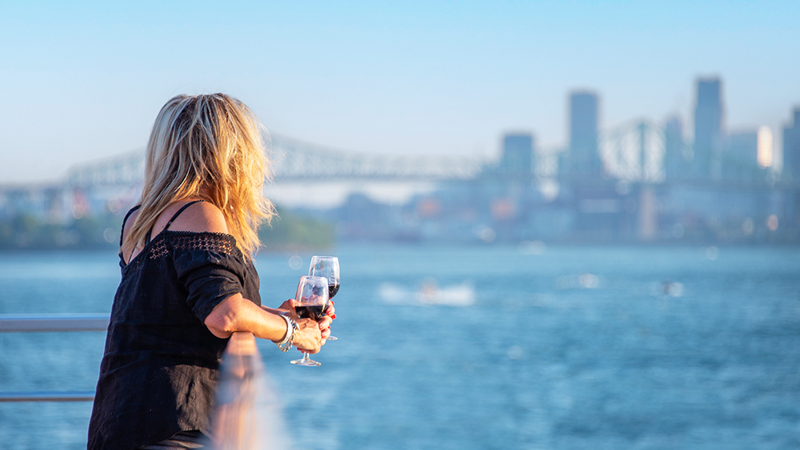 A woman holding a glass of red wine on one of the terraces of the AML Cavalier Maxim ship sailing on the St. Lawrence River, with a view of the Montreal skyline in the background.