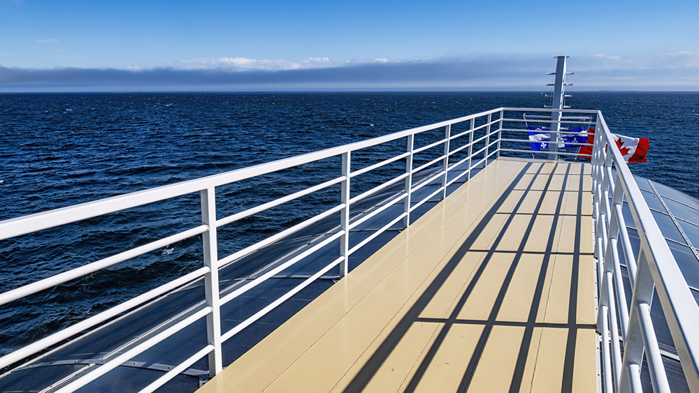 A view of the upper deck of a boat, lined with white railings, providing a clear perspective of the vast blue sea under a clear sky. A Canadian flag flutters in the background, gently swayed by the wind.