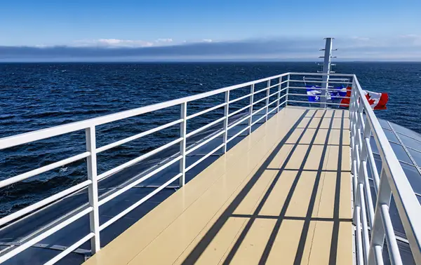 A view of the upper deck of a boat, lined with white railings, providing a clear perspective of the vast blue sea under a clear sky. A Canadian flag flutters in the background, gently swayed by the wind.