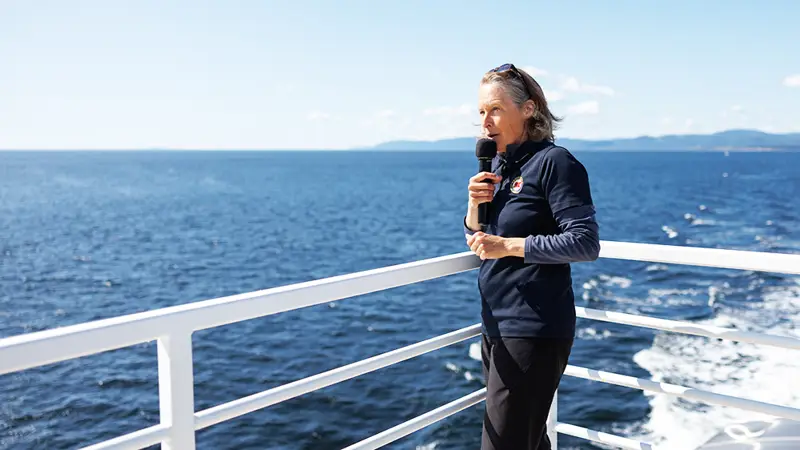 A naturalist stands on a boat's deck holding a microphone, overlooking the vast ocean under a clear blue sky. Distant landforms are visible across the water. The person wears a dark outfit and appears to be speaking.