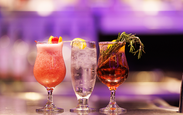 Trois cocktails de couleurs et de garnitures variées sur un comptoir de bar, avec un fond légèrement flou mettant en valeur les boissons vibrantes.