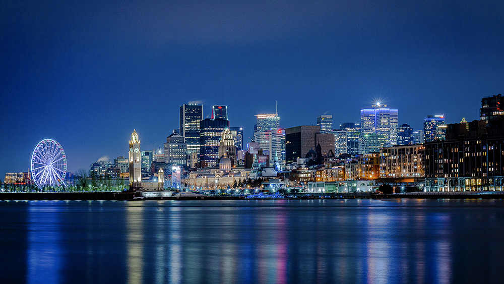 Nighttime view of a city illuminated with skyscrapers, a lit-up Ferris wheel, and reflections on the calm water in the foreground.