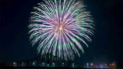 A colorful firework, with shades of green and purple, bursts in the night sky above an iconic bridge, creating a spectacular view.