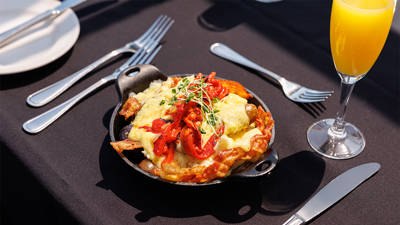 A hot dish served in a small cast iron skillet, topped with grilled vegetables, melted cheese, and fresh sprouts, accompanied by a glass of mimosa on an elegantly set table.