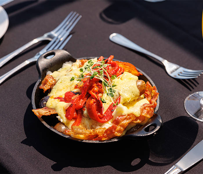 A hot dish served in a small cast iron skillet, topped with grilled vegetables, melted cheese, and fresh sprouts, accompanied by a glass of mimosa on an elegantly set table.