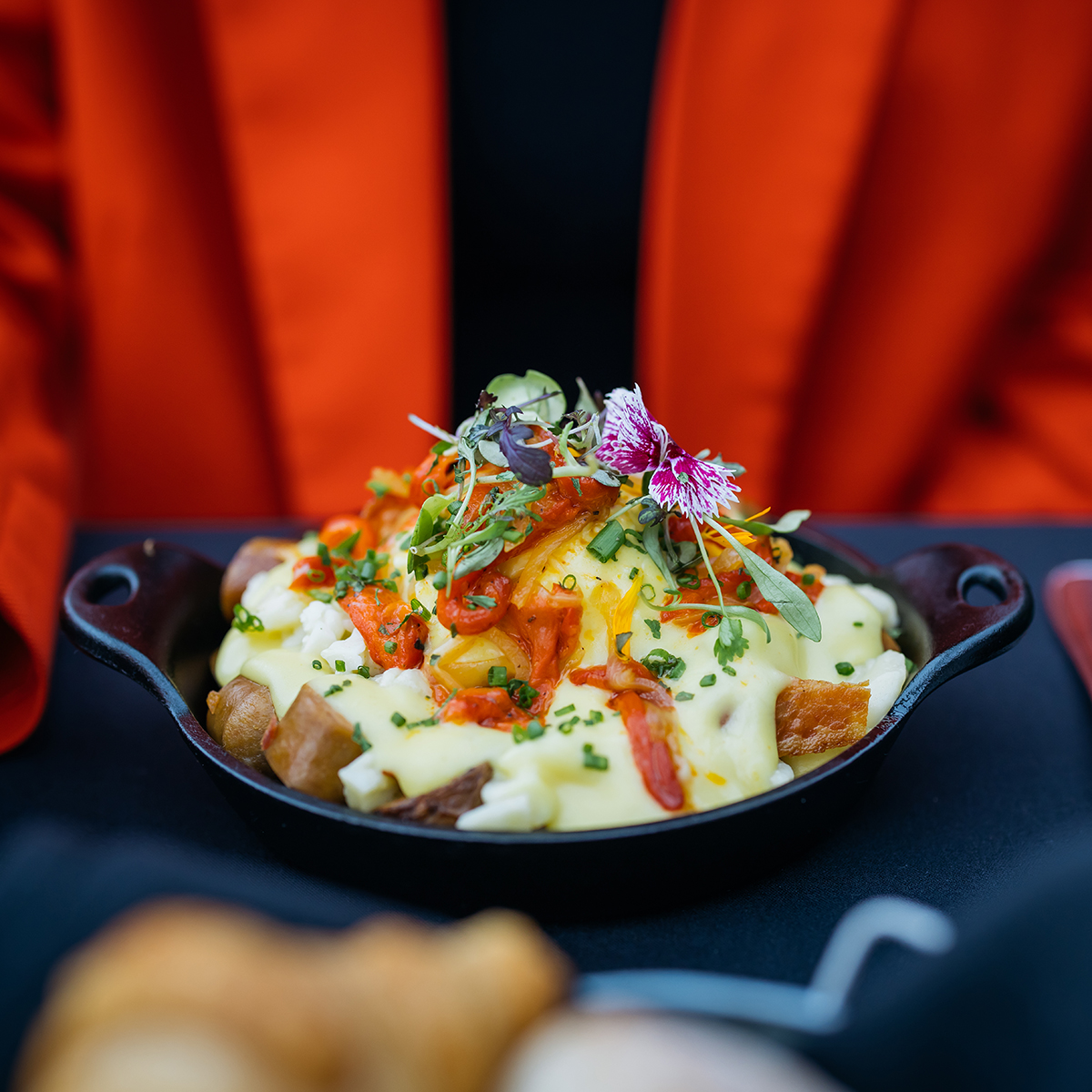 Un plat gourmand composé de pommes de terre rôties, nappées de sauce hollandaise, garnies de légumes, herbes fraîches et fleurs comestibles, servi dans une poêle en fonte.