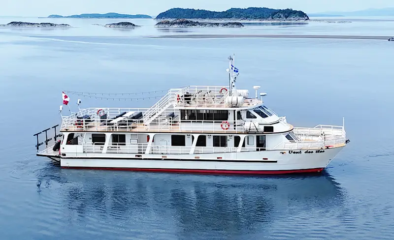 Le bateau "Vent des Îles" navigue tranquillement sur des eaux calmes entourées d'îles et d'une légère brume à l'horizon. Le ciel est dégagé et lumineux, créant une ambiance paisible.