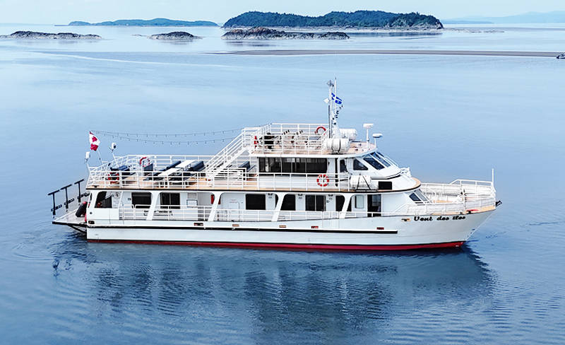 The ship "Vent des Îles" sails peacefully on calm waters surrounded by islands and a light mist on the horizon. The sky is clear and bright, creating a serene atmosphere.