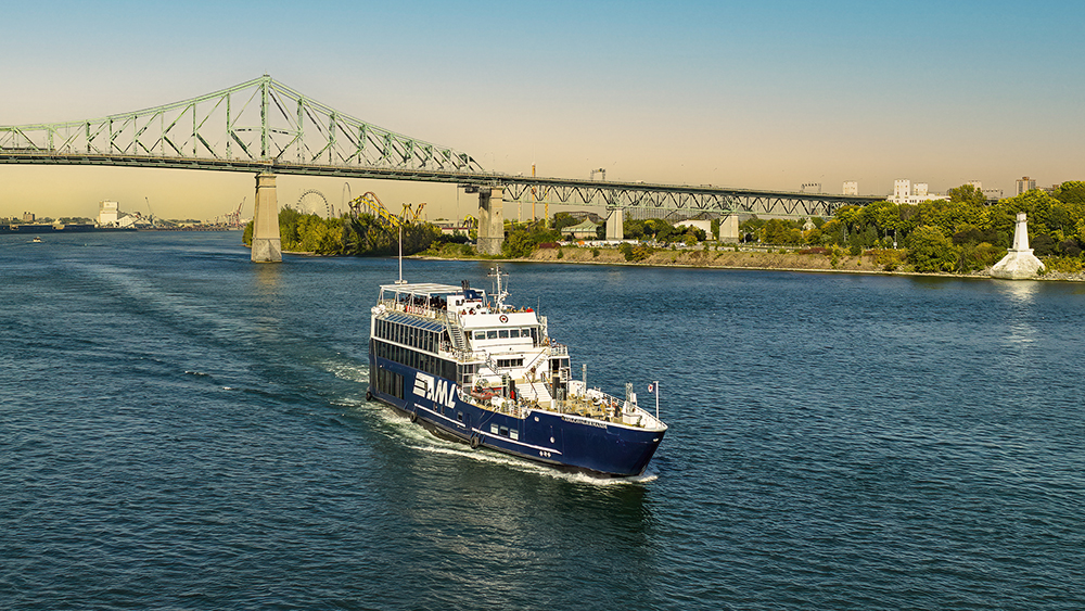 Un bateau de croisière bleu navigue sur une rivière par une journée ensoleillée, avec le pont Jacques-Cartier et des arbres en arrière-plan.