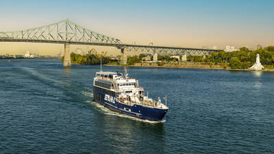 A blue cruise ship sails on a river on a sunny day, with the Jacques-Cartier Bridge and a green shoreline in the background.