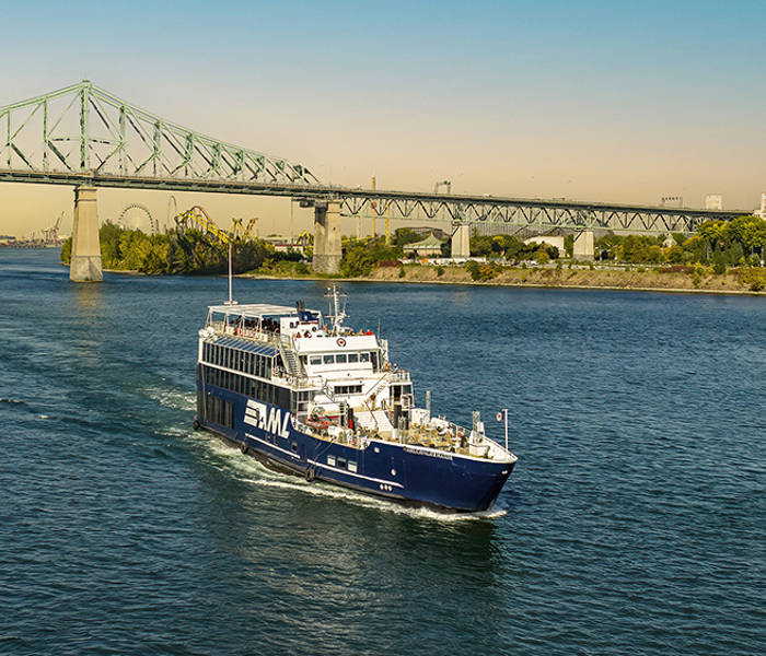 A blue cruise ship sails on a river on a sunny day, with the Jacques-Cartier Bridge and a green shoreline in the background.