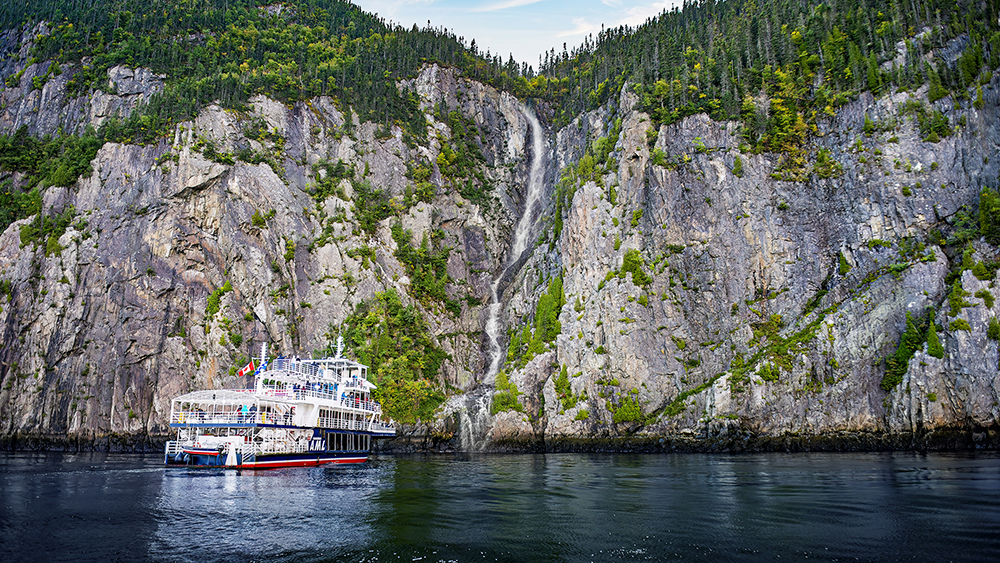 Un bateau de croisière est amarré près d'une imposante falaise rocheuse recouverte de végétation. Une cascade étroite descend le long de la falaise pour se jeter dans l'eau en contrebas, sous un ciel bleu avec quelques nuages.