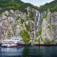 A cruise ship is docked near a towering rocky cliff covered in vegetation. A narrow waterfall flows down the cliff, cascading into the water below, under a blue sky with a few clouds.