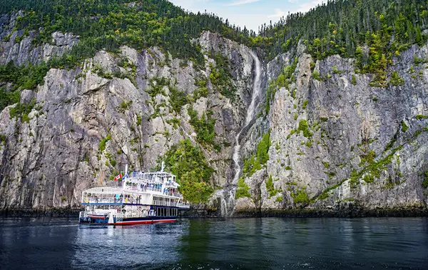 A cruise ship is docked near a towering rocky cliff covered in vegetation. A narrow waterfall flows down the cliff, cascading into the water below, under a blue sky with a few clouds.