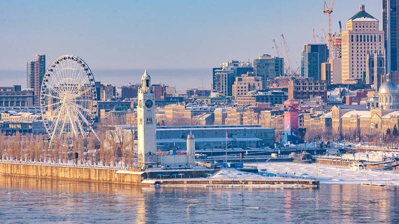 Panoramic view of Old Port of Montreal in winter, featuring the Clock Tower, the Ferris wheel, and downtown buildings under a clear sky.