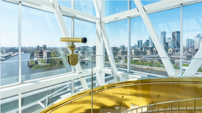 A telescope in a modern observatory with panoramic windows overlooking the Montreal skyline under a blue sky.