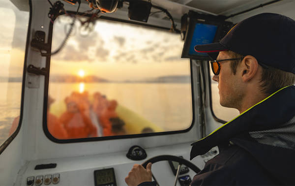 A captain wearing a cap and sunglasses steers a boat while looking through the windshield. Outside, a sunset over the water is visible, with blurred passengers at the back of the boat. The sky is partly cloudy, creating a warm atmosphere.