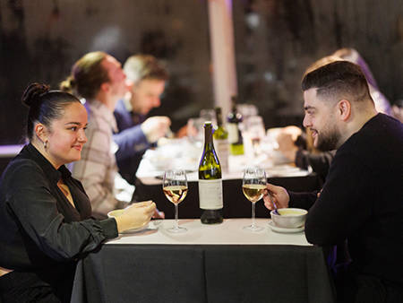 A couple shares a meal in an elegant setting, with two glasses of white wine and a bottle on the table. Other diners are visible in the background.