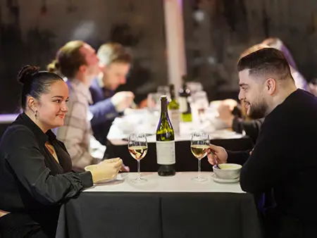 A couple shares a meal in an elegant setting, with two glasses of white wine and a bottle on the table. Other diners are visible in the background.
