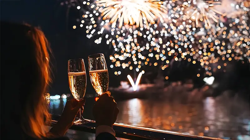 Deux personnes trinquent avec des verres de champagne sur un balcon, admirant un feu d'artifice spectaculaire illuminant le ciel nocturne et se reflétant sur l'eau.