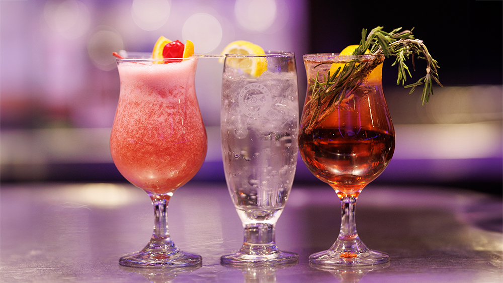 Three elegant cocktails lined up on a counter, featuring a pink cocktail garnished with a cherry, a clear drink with a lemon slice, and an amber cocktail decorated with rosemary.