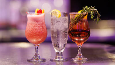 Three elegant cocktails lined up on a counter, featuring a pink cocktail garnished with a cherry, a clear drink with a lemon slice, and an amber cocktail decorated with rosemary.